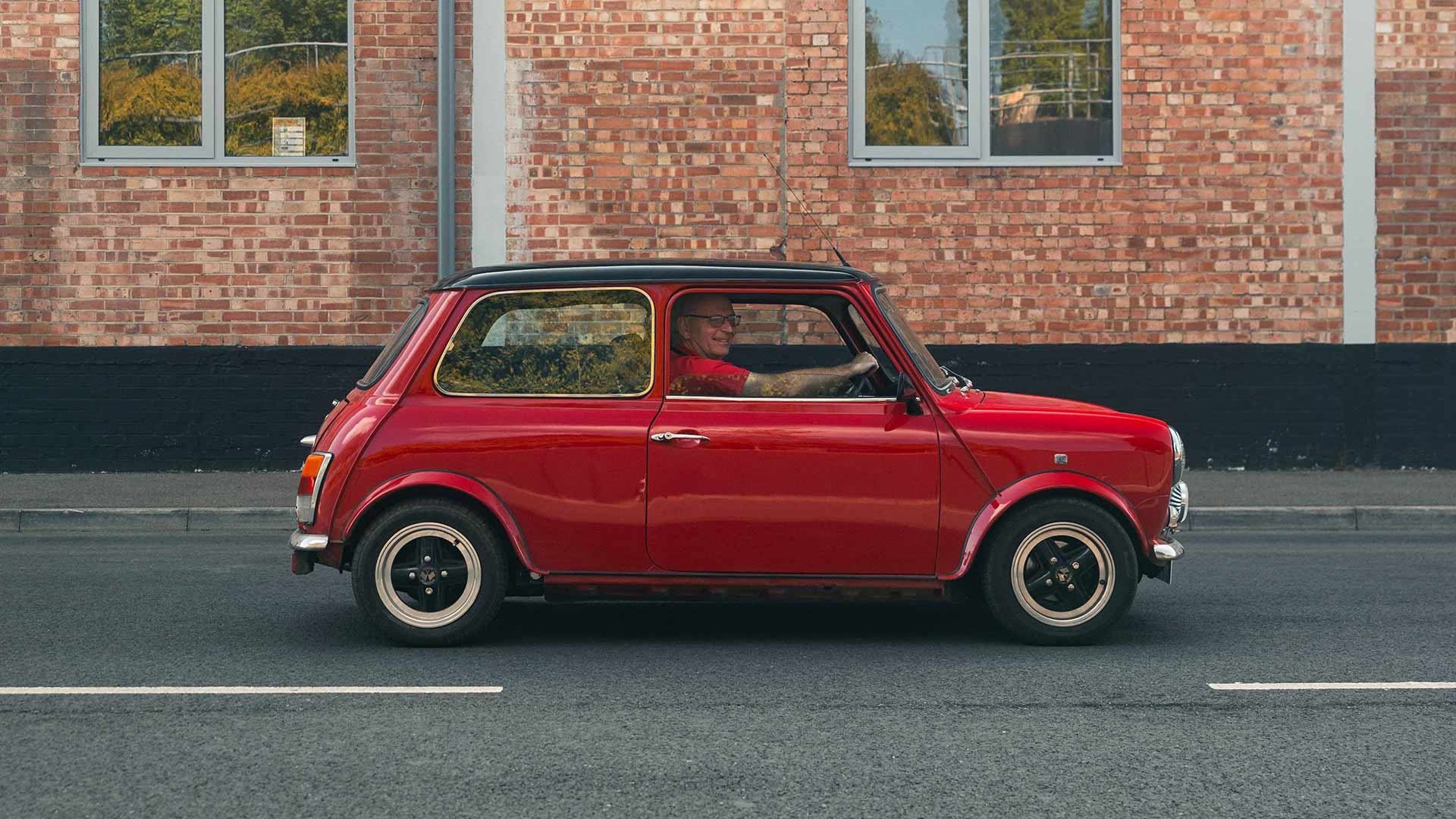 A red vintage Mini Cooper drives past a brick wall on a sunny day. The driver is visible, windows show reflections of trees, creating a nostalgic feel.